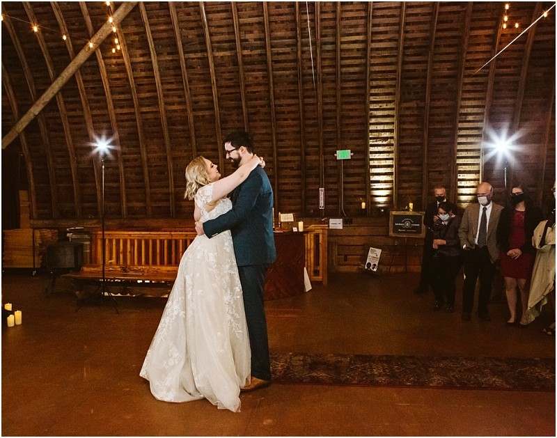 A bride and groom share their first dance in a rustic snohomish barn with wooden walls and string lights while a small group of guests watches from the side Snohomish wedding photography