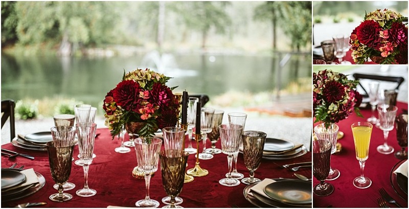 A formal table setting with wine glasses glassware and dark plates on a red tablecloth featuring a centerpiece of red and green flowers set outdoors by a lake in snohomish Snohomish wedding photography