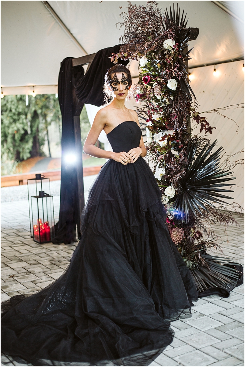 A woman in a strapless black ball gown and black lace mask stands under a floral arch with dark leaves and white flowers at an elegant snohomish outdoor event with string lights and a lantern with red candles nearby Snohomish wedding photography