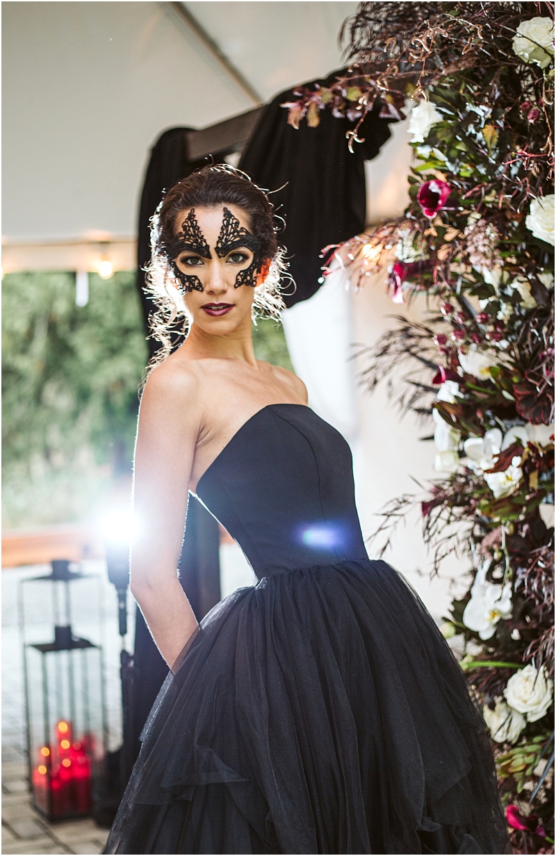 A woman in a strapless black gown and dramatic black and gold mask stands beside a snohomish inspired floral arrangement with white and dark red flowers illuminated by bright light from behind Snohomish wedding photography