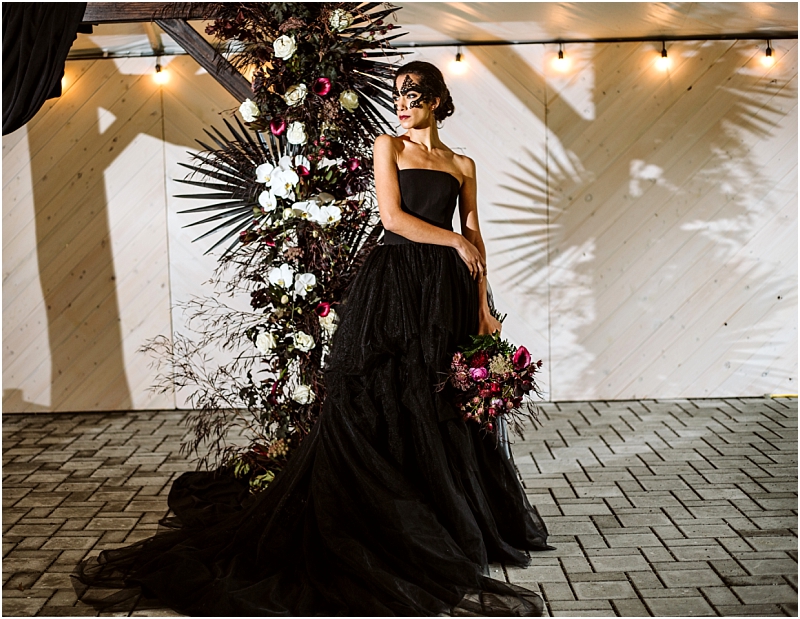 A woman in a strapless black gown and black mask stands by a dramatic floral arrangement with dark and white flowers holding a bouquet under string lights on a paved floor at an elegant snohomish venue Snohomish wedding photography