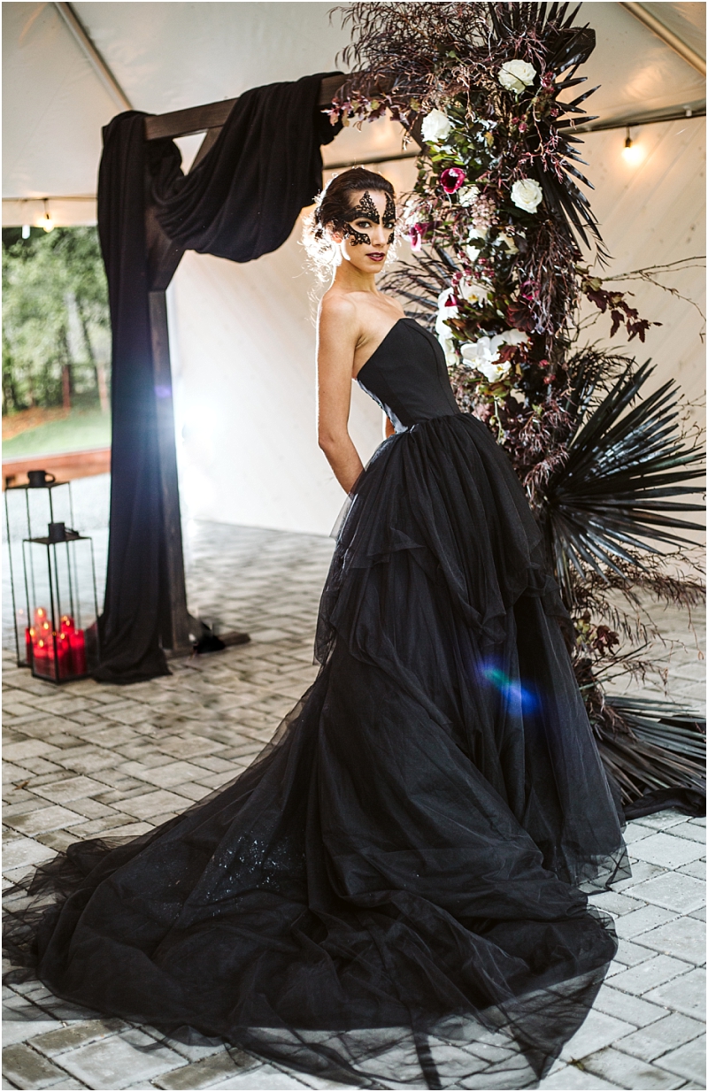 A woman in a black ball gown and black lace mask poses beside a dramatic floral arrangement with dark leaves and white roses at a snohomish event under a tent decorated with black drapery and red candles Snohomish wedding photography