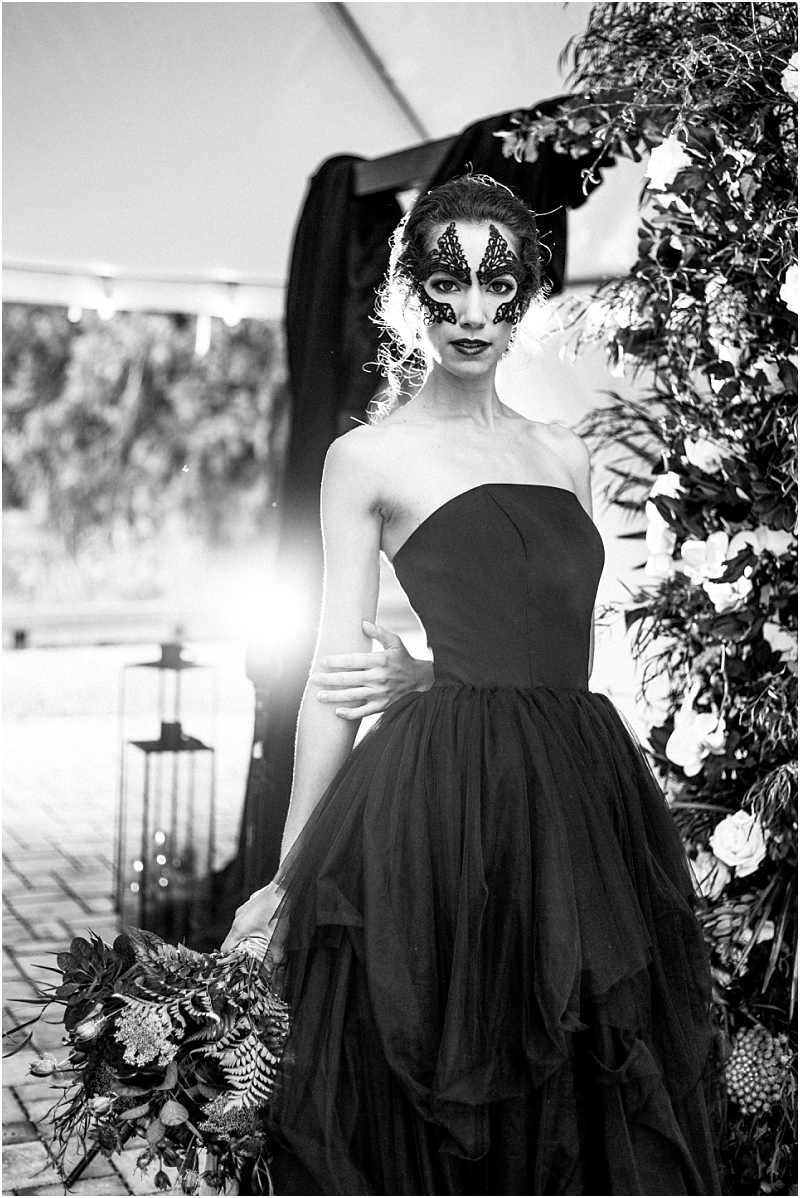 A woman in a strapless black gown holds a bouquet and wears dramatic mask like face makeup She stands near floral arrangements in snohomish with sunlight shining behind her The photo is in striking black and white Snohomish wedding photography