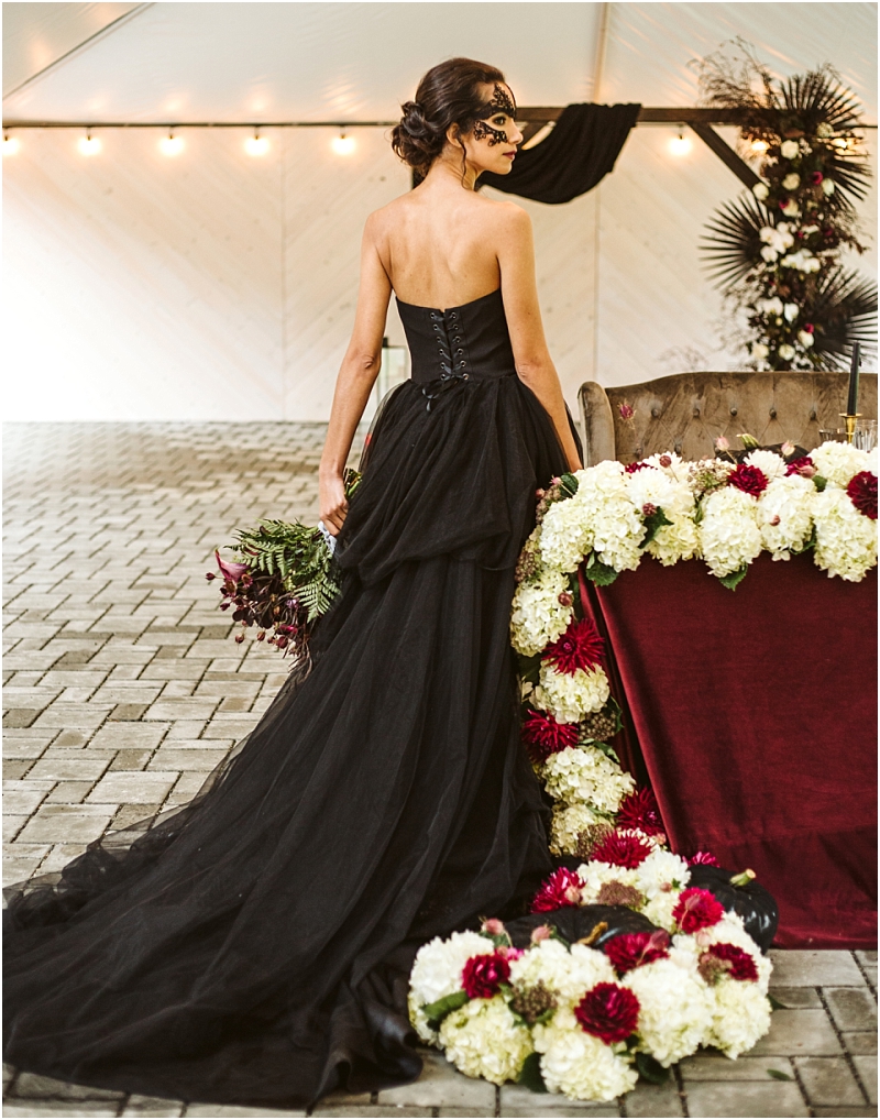 A woman in a strapless black gown and black lace mask stands beside a table decorated with white and dark red flowers and a burgundy tablecloth holding a bouquet beneath romantic string lights at a snohomish venue Snohomish wedding photography