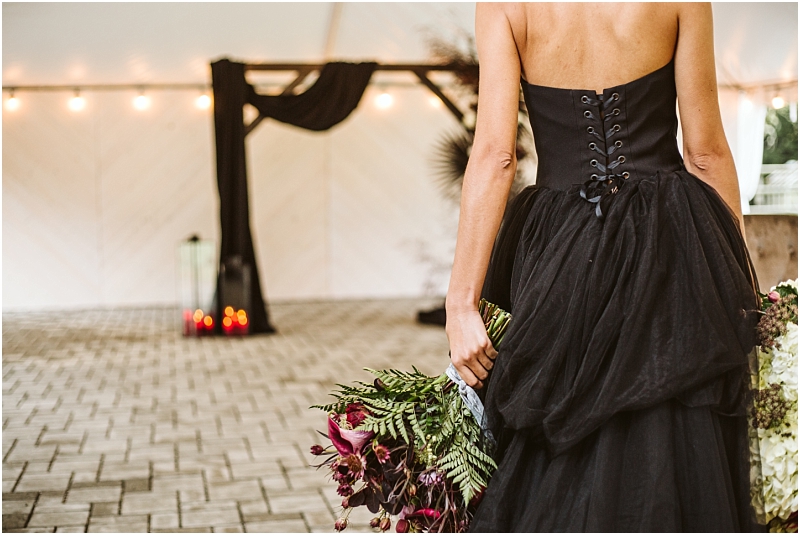 A woman in a strapless black dress holds a bouquet of dark purple flowers standing outdoors in front of a black draped wedding arch with candles in the background at a romantic snohomish venue Snohomish wedding photography