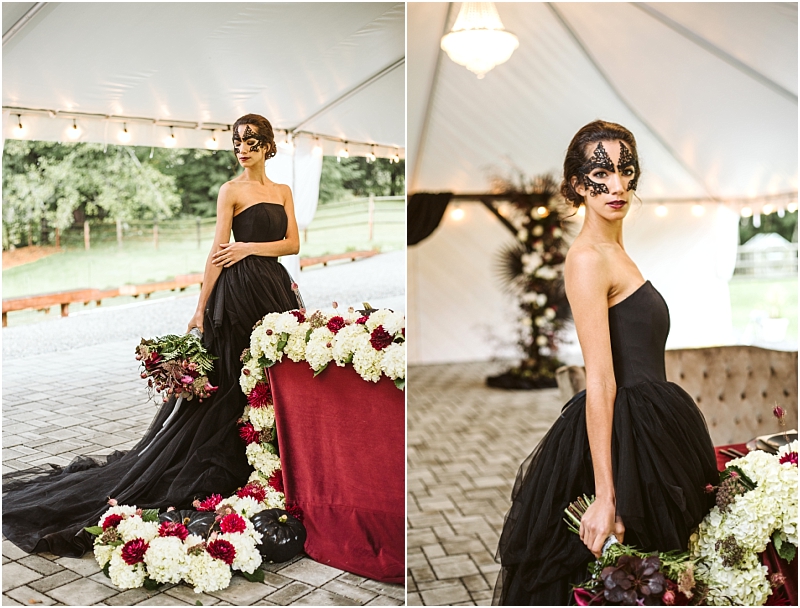 A woman in a strapless black gown and lace mask stands under a white tent in snohomish holding a bouquet of dark flowers She is surrounded by red and white floral arrangements on a table beside her Snohomish wedding photography