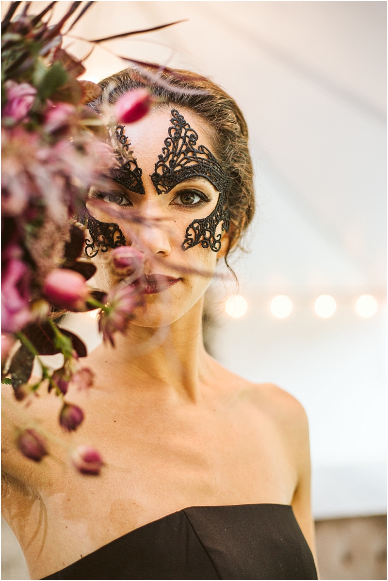A woman with elaborate black lace face paint looks at the camera wearing a strapless black dress She stands beside a bouquet of dark flowers with snohomishs soft lights blurred in the background Snohomish wedding photography
