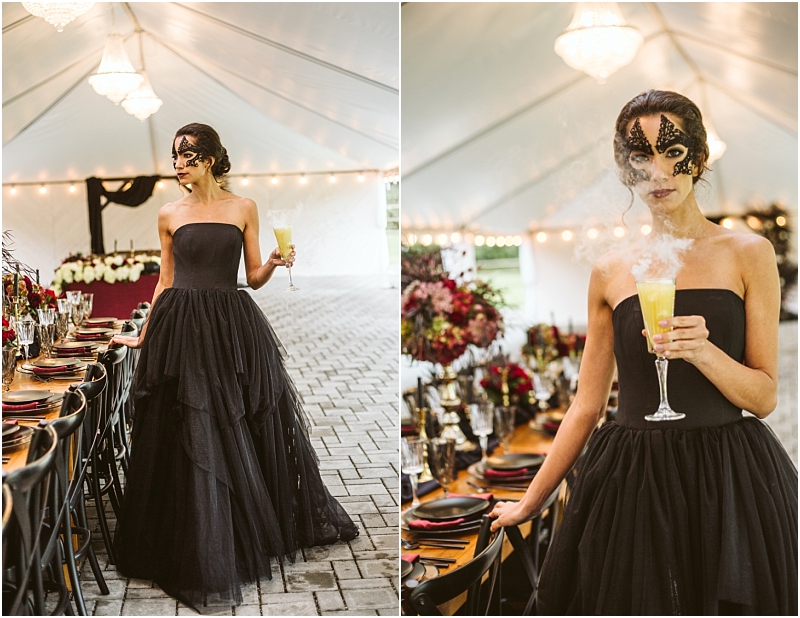 A woman in a black strapless gown and ornate black mask holds a smoking yellow cocktail standing by an elegantly set table under a white tent with string lights and chandeliers at a chic snohomish event Snohomish wedding photography