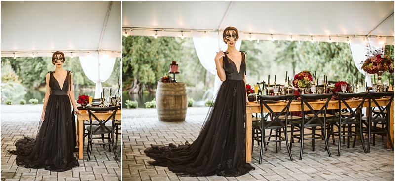 A woman in a flowing black gown and lace mask stands beside an elegantly set wooden table under a white canopy with lush greenery and floral centerpieces in the background of this enchanting snohomish setting Snohomish wedding photography