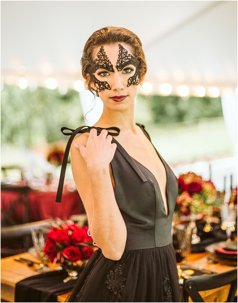 A woman in a black deep v neck dress and an ornate black lace mask stands by a table set with red flowers and elegant glassware at a formal snohomish event under a tent Snohomish wedding photography