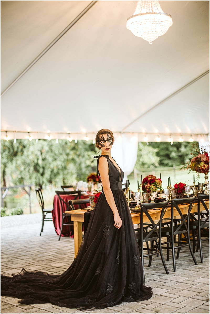 A woman in a dramatic black gown and ornate black mask stands under a tented chandelier at a snohomish outdoor event beside an elegantly set table with red floral arrangements and black chairs Snohomish wedding photography