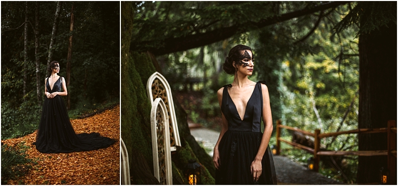 A woman in a deep v neck black gown and lace mask stands in a forested snohomish area surrounded by tall trees moss and a rustic wooden fence She gazes thoughtfully into the distance Snohomish wedding photography