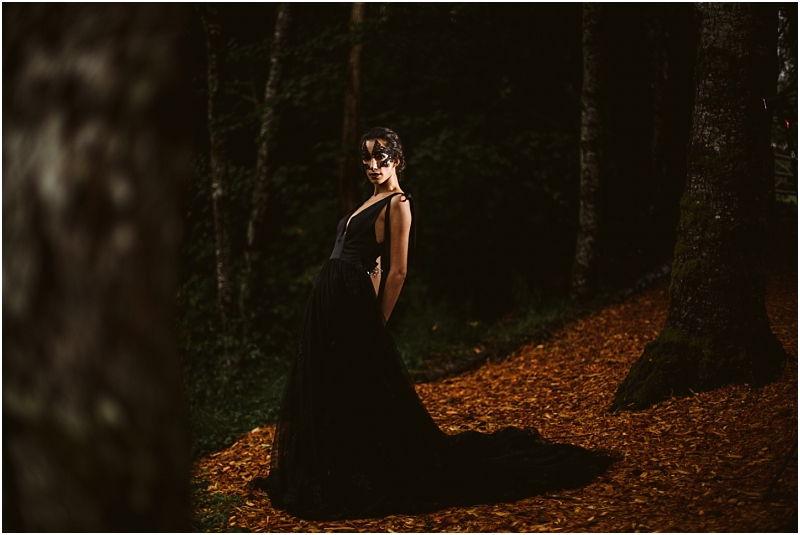 A woman in a long elegant black dress stands on a leaf covered path in a dark snohomish forest looking over her shoulder as dramatic lighting highlights her figure against the shadowy trees Snohomish wedding photography
