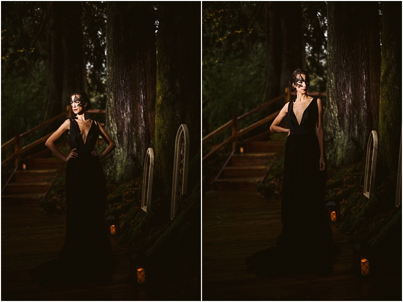 A woman in a long black deep v neck gown and a decorative black mask stands on a wooden pathway in a snohomish forest at night with lanterns and framed mirrors propped against tree trunks nearby Snohomish wedding photography