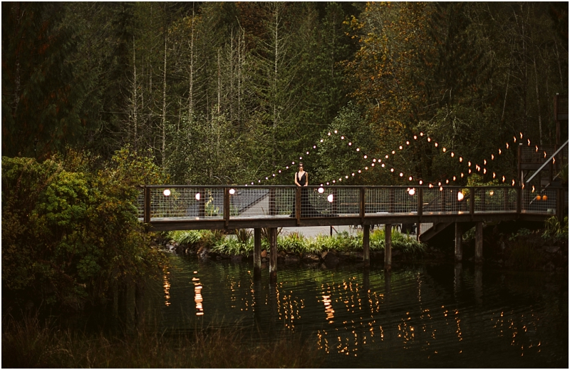 A person stands alone on a wooden bridge decorated with string lights over a calm reflective body of water surrounded by green forest and trees in scenic snohomish Snohomish wedding photography