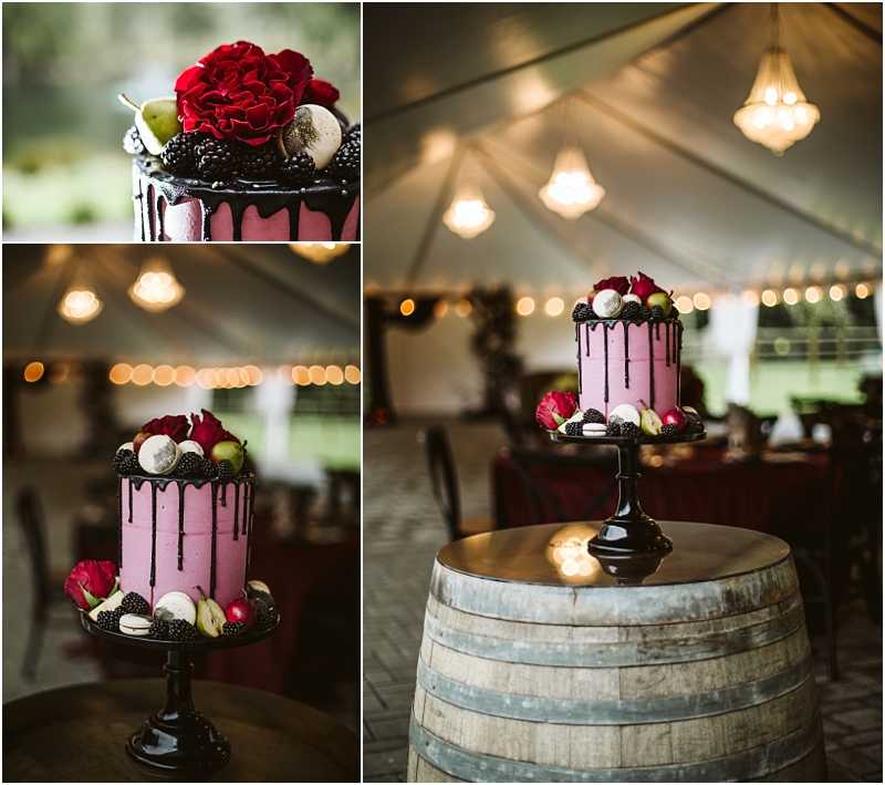 A pink drip cake decorated with berries macarons and red flowers sits on a black cake stand atop a wooden barrel at a snohomish event displayed under a tent with chandeliers and string lights in the background Snohomish wedding photography