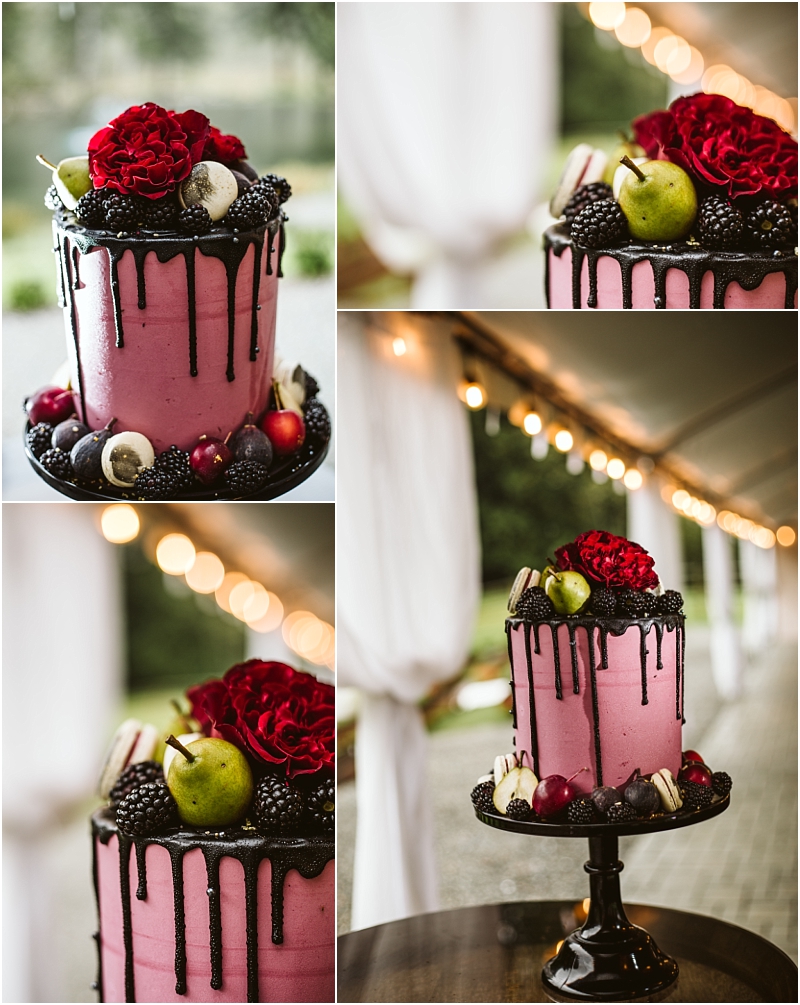A tall pink cake with dark chocolate drip topped with red roses figs pears blackberries and plums is displayed on a black cake stand at an outdoor snohomish event under a white tent Snohomish wedding photography