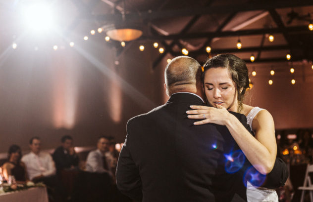 A bride in a white dress hugs an older man in a suit likely her father while dancing at a warmly lit snohomish wedding reception Guests are seated in the background under glowing string lights Snohomish wedding photography