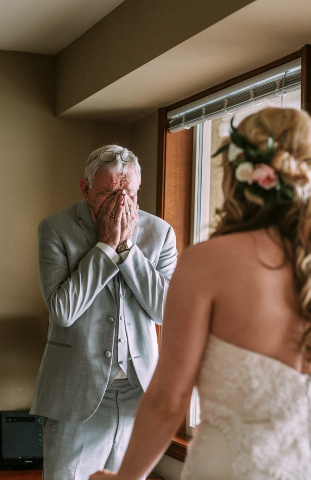 An older man in a light gray suit covers his face with his hands appearing emotional while facing a woman in a white strapless dress with flowers in her hair near a sunlit window in snohomish Snohomish wedding photography