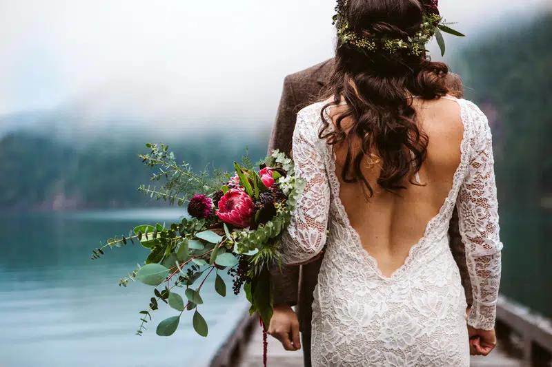 A bride in a lace, open-back dress holding a bouquet of flowers stands on a dock by a misty Snohomish lake, facing a person in a brown suit. Forested hills are visible in the foggy background. Snohomish Wedding Photography