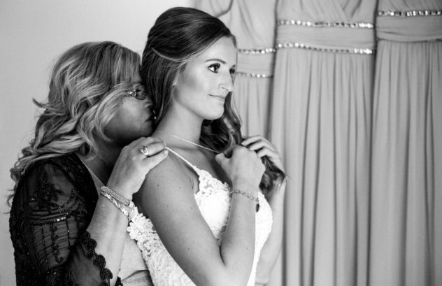 A woman helps a bride adjust her necklace as they prepare for a snohomish wedding The bride wears a white dress emotion fills the air and tender moments are shared as bridesmaid dresses hang in the background Snohomish wedding photography