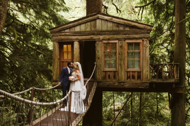 A bride and groom stand on a rope bridge leading to a rustic wooden treehouse in a lush snohomish forest The bride holds a bouquet while the couple embraces surrounded by tall trees and vibrant greenery Snohomish wedding photography Washington wedding venue treehouse point