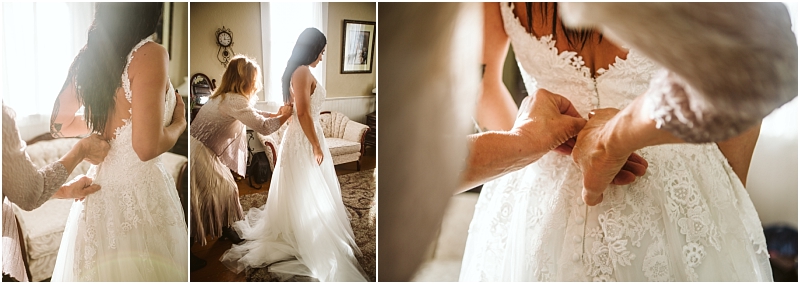 A bride in a lace wedding dress is being helped by a woman to fasten the back of her gown The softly lit snohomish room showcases close up and wider views of the dress and the supportive hands at work Snohomish wedding photography