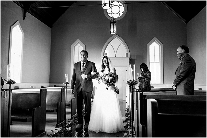 A bride in a white gown walks down the aisle with a man in a suit inside a small snohomish church Two people stand watching on either side sunlight streams through tall windows creating a bright serene atmosphere Snohomish wedding photography
