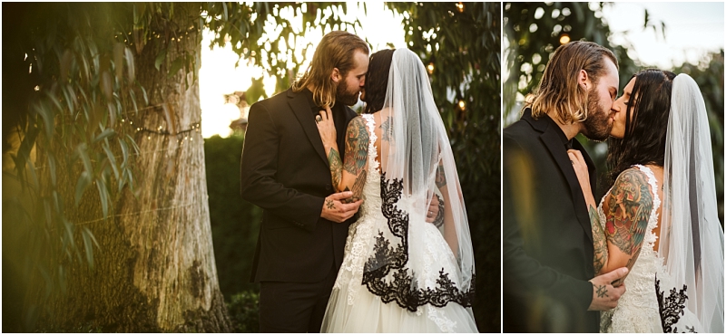 A bride in a white dress with a black lace veil and a groom in a black suit share a romantic kiss outdoors near a tree surrounded by snohomish greenery at sunset Snohomish wedding photography