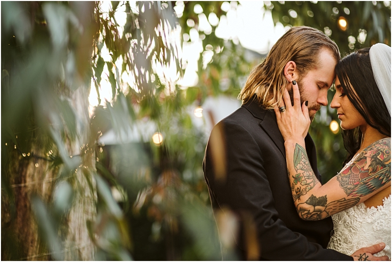 A couple embraces closely outdoors in snohomish surrounded by greenery The woman with dark hair and colorful tattoos gently touches the mans face Both appear emotional and connected dressed in wedding attire as sunlight filters through the leaves Snohomish wedding photography
