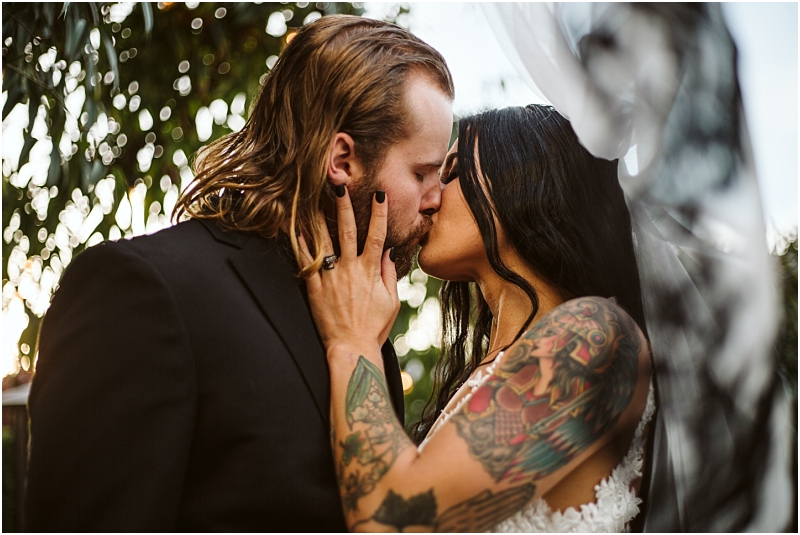 A couple kisses passionately outdoors in snohomish the man wears a black suit and has long hair while the woman in a white dress has long dark hair and colorful tattoos Sunlight filters through greenery in the background Snohomish wedding photography