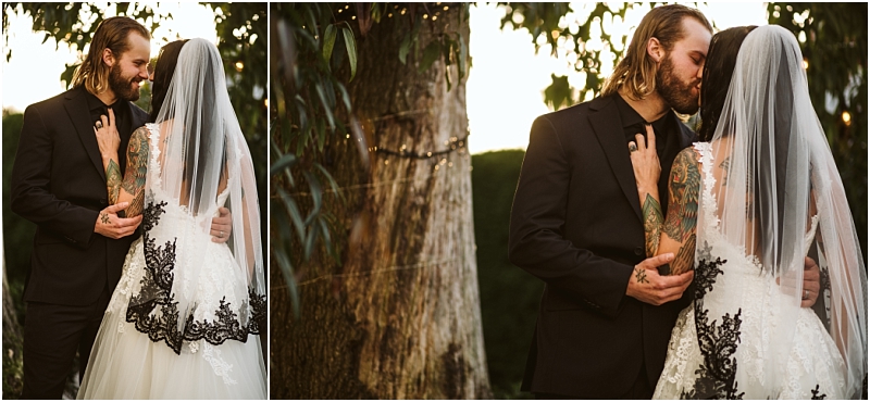 A bride and groom embrace and kiss outdoors by a tree in snohomish The bride wears a white dress with black lace and a veil while the groom dons a black suit String lights are wrapped around the tree in the background Snohomish wedding photography