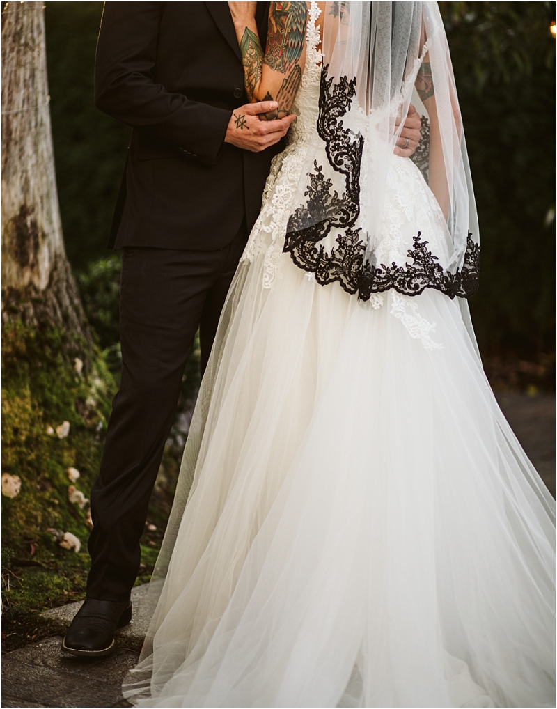 A bride in a white gown with black lace trim on her veil embraces a groom in a black suit Both have visible tattoos as they stand outdoors beside a tree in snohomish Faces are not visible Snohomish wedding photography
