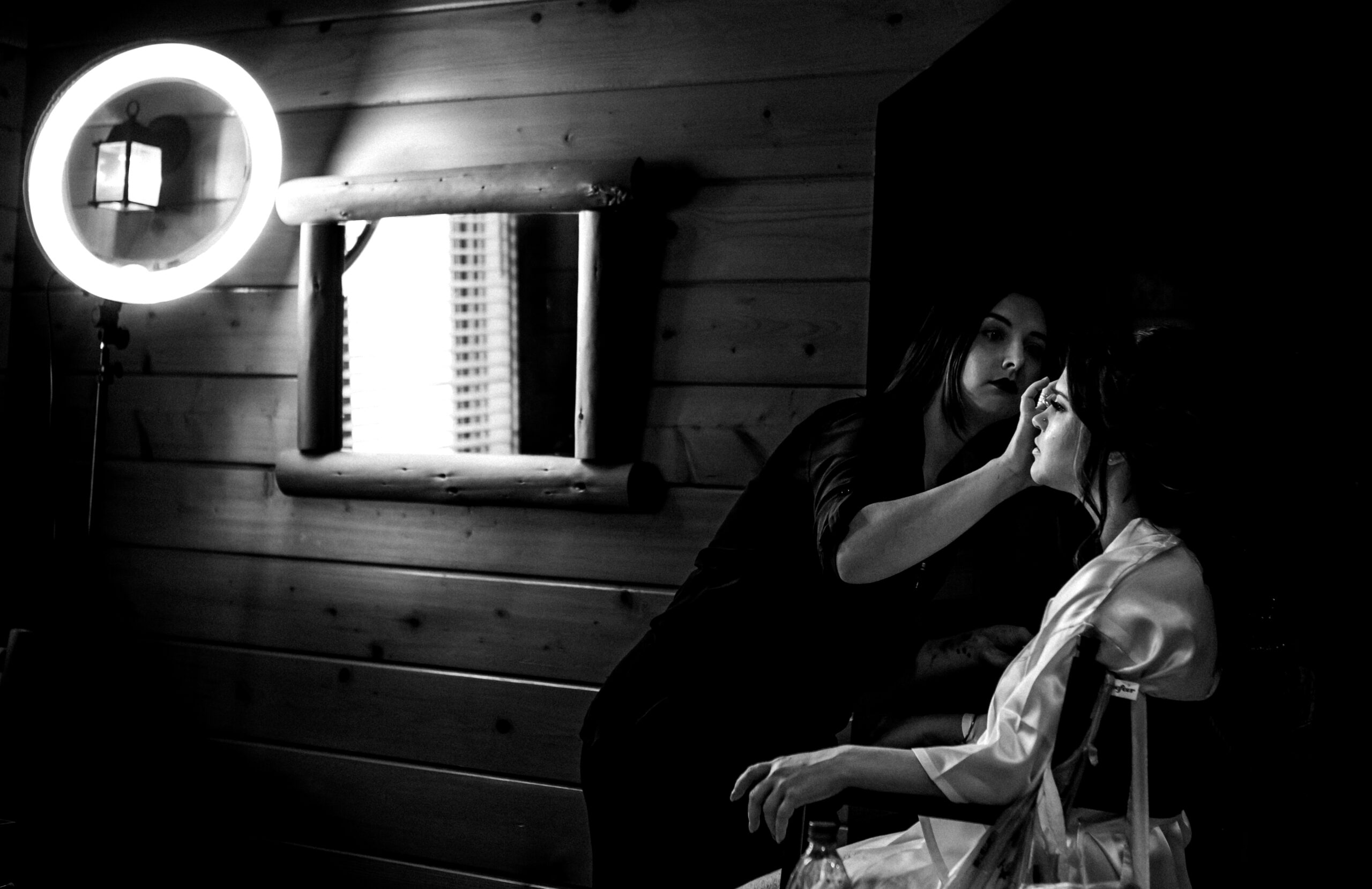 A woman in a robe sits while another woman applies her makeup in a dimly lit Snohomish room with wooden walls; a round ring light and a window with blinds are visible in the background. Snohomish Wedding Photography