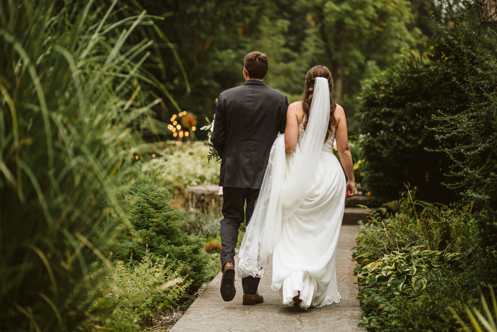 A bride and groom walk hand in hand down a lush Snohomish garden path. The bride's white dress and flowing veil trail behind, while the groom, dressed in a dark suit, strolls by her side. They are photographed from behind amidst vibrant greenery. Snohomish Wedding Photography