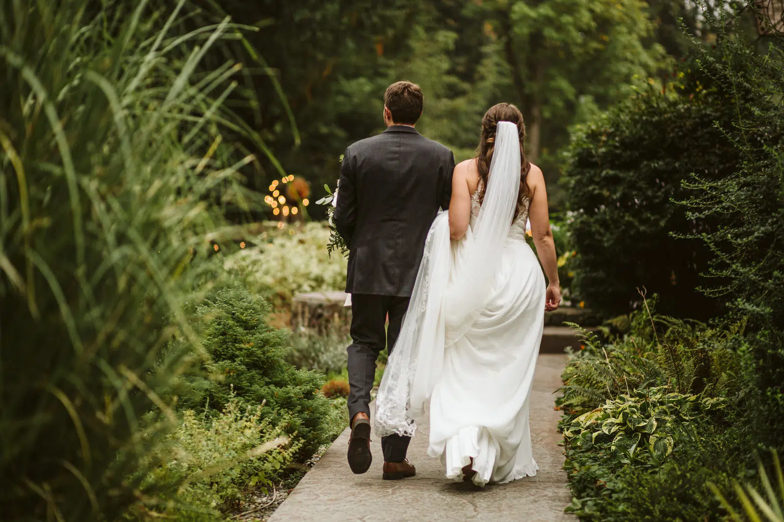 A bride and groom walk hand in hand down a lush Snohomish garden path. The bride's white dress and flowing veil trail behind, while the groom, dressed in a dark suit, strolls by her side. They are photographed from behind amidst vibrant greenery. Snohomish Wedding Photography