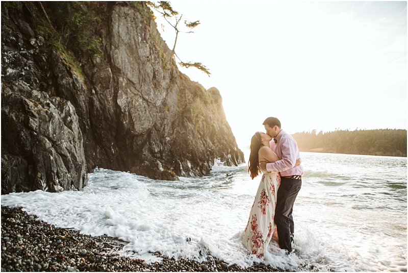 A couple stands in the surf on a rocky snohomish beach at sunset embracing and kissing as waves wash around them The woman wears a floral dress the man a light shirt and dark pants Cliffs and trees rise nearby Snohomish wedding photography A couple stands in the surf on a rocky snohomish beach at sunset embracing and kissing as waves wash around them The woman wears a floral dress the man a light shirt and dark pants Cliffs and trees rise nearby Snohomish wedding photography