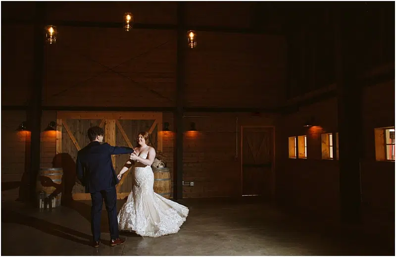 A bride and groom dance together in a dimly lit Snohomish rustic barn with wooden walls, barrels, and soft warm lighting overhead. Snohomish Wedding Photography