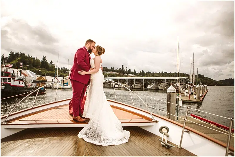 A bride in a white lace gown and a groom in a burgundy suit embrace on the bow of a boat in Snohomish, with the marina, sparkling water, and cloudy sky creating a romantic backdrop. Snohomish Wedding Photography