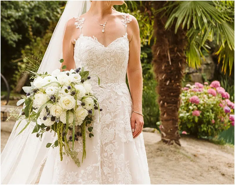 A bride in an elegant lace wedding dress holds a large bouquet of white roses and greenery, standing outdoors near palm trees and flowering bushes at a scenic Snohomish venue. Her face is not visible. Snohomish Wedding Photography