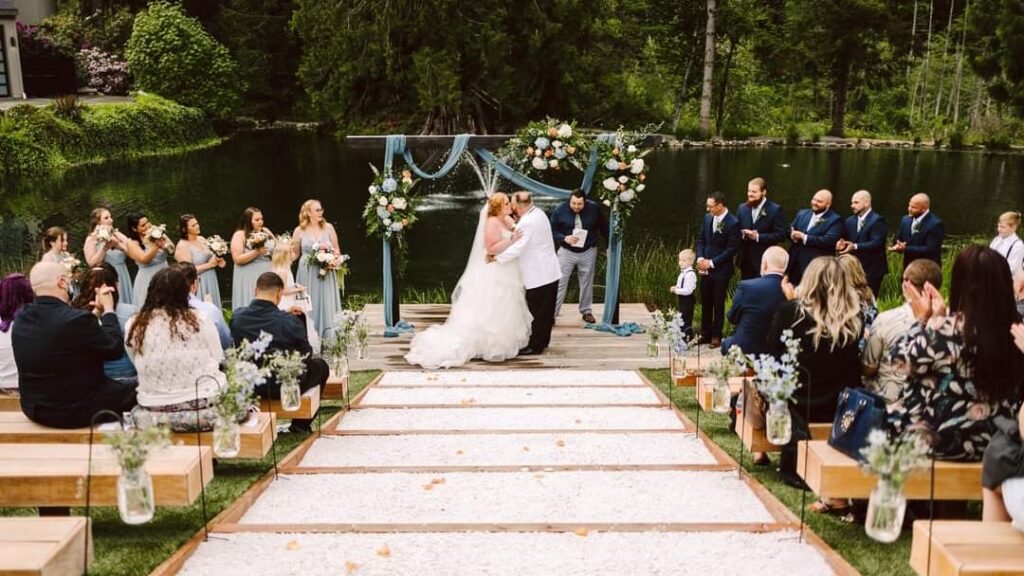 A bride and groom kiss at an outdoor lakeside wedding ceremonyone of the most picturesque Seattle wedding venuessurrounded by their wedding party seated guests on wooden benches floral decorations and lush greenery in the background Snohomish Wedding Photography A bride and groom kiss at an outdoor lakeside wedding ceremony—one of the most picturesque Seattle wedding venues—surrounded by their wedding party, seated guests on wooden benches, floral decorations, and lush greenery in the background. Snohomish Wedding Photography