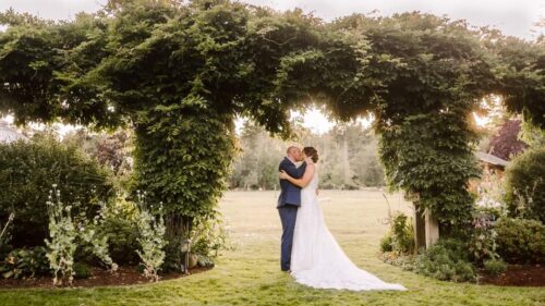 A bride and groom embrace and kiss under a large leafy green arch in a garden setting at one of the enchanting Washington wedding venues with sunlight filtering through the trees in the background Snohomish Wedding Photography A bride and groom embrace and kiss under a large, leafy green arch in a garden setting at one of the enchanting Washington wedding venues, with sunlight filtering through the trees in the background. Snohomish Wedding Photography