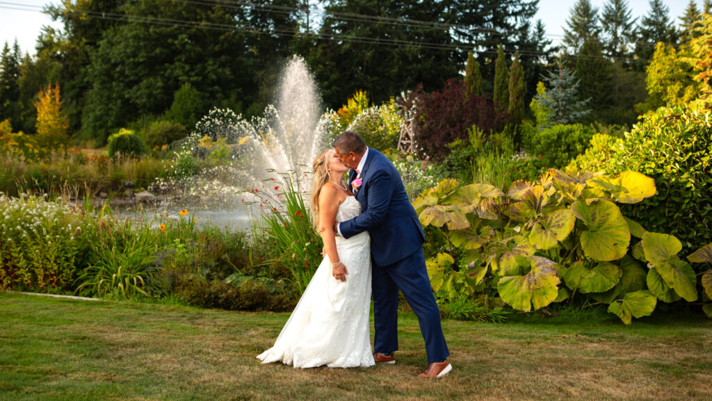A bride and groom share a kiss outdoors on a lush green lawn at one of the picturesque Snohomish wedding venues with a fountain and vibrant garden plants in the background on a sunny day Snohomish Wedding Photography A bride and groom share a kiss outdoors on a lush green lawn at one of the picturesque Snohomish wedding venues, with a fountain and vibrant garden plants in the background on a sunny day. Snohomish Wedding Photography