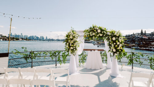 A wedding arch decorated with white flowers and greenery stands on a boat deck overlooking a city skylineone of the most unique Washington wedding venueswhite chairs await guests with sparkling water in the background under a sunny sky Snohomish Wedding Photography A wedding arch decorated with white flowers and greenery stands on a boat deck overlooking a city skyline—one of the most unique Washington wedding venues—white chairs await guests, with sparkling water in the background under a sunny sky. Snohomish Wedding Photography