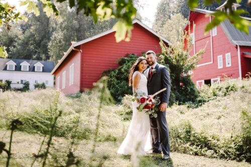 A bride and groom stand together outdoors in front of a large red barn surrounded by greenery Perfect for Washington weddings the couple smiles in wedding attire under natural sunlight as the bride holds a bouquet of flowers Snohomish Wedding Photography A bride and groom stand together outdoors in front of a large red barn, surrounded by greenery. Perfect for Washington weddings, the couple smiles in wedding attire under natural sunlight as the bride holds a bouquet of flowers. Snohomish Wedding Photography