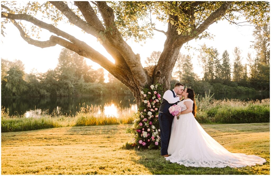 Summer pemberton farm wedding with floral tree wrap and bride in white gown with bouquet kissing groom in blue suit in the golden sunset by the snohomish river Summer pemberton farm wedding with floral tree wrap and bride in white gown with bouquet kissing groom in blue suit in the golden sunset by the snohomish river