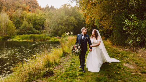 A bride and groom walk hand in hand along a grassy path beside a pond at one of the charming Snohomish wedding venues surrounded by lush green trees and autumn foliage The bride holds her dress and bouquet as they smile warmly at each other Snohomish Wedding Photography A bride and groom walk hand in hand along a grassy path beside a pond at one of the charming Snohomish wedding venues, surrounded by lush green trees and autumn foliage. The bride holds her dress and bouquet as they smile warmly at each other. Snohomish Wedding Photography