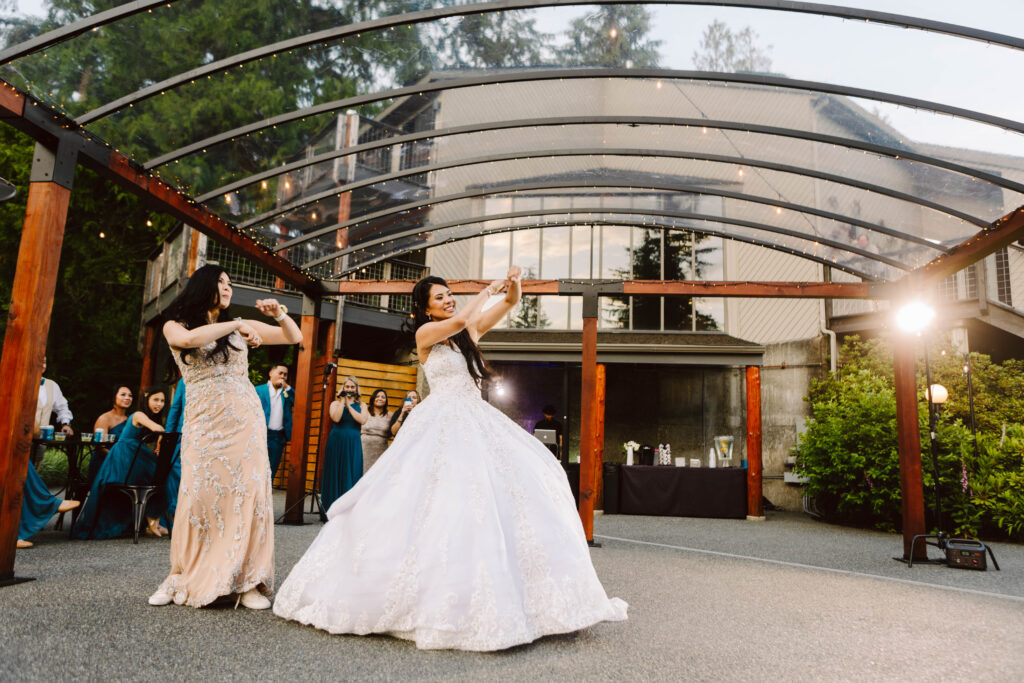 Snohomish Wedding Photographer GSquared Weddings Two women in elegant gowns dance under a transparent canopy with wooden beams. The woman in white, likely the bride, shares this joyous wedding moment with her mom. Guests in formal attire stand smiling and capturing photos at this lively outdoor celebration. Serving Seattle, Snohomish and Washington State