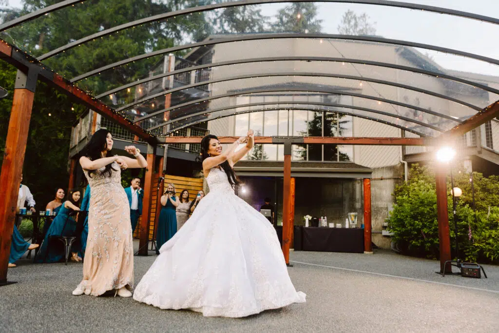 Snohomish Wedding Photographer GSquared Weddings Two women in elegant gowns dance under a transparent canopy with wooden beams The woman in white likely the bride shares this joyous wedding moment with her mom Guests in formal attire stand smiling and capturing photos at this lively outdoor celebration Serving Seattle Snohomish and Washington State Snohomish Wedding Photographer GSquared Weddings Two women in elegant gowns dance under a transparent canopy with wooden beams. The woman in white, likely the bride, shares this joyous wedding moment with her mom. Guests in formal attire stand smiling and capturing photos at this lively outdoor celebration. Serving Seattle, Snohomish and Washington State