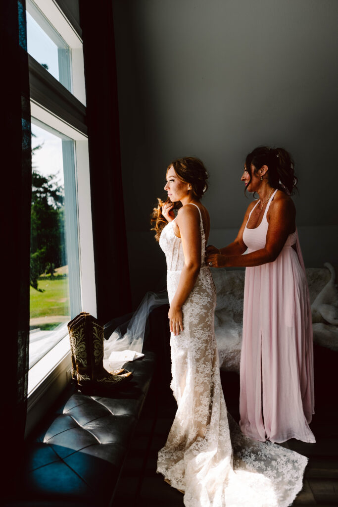 Snohomish Wedding Photographer GSquared Weddings A bride in a lace gown stands near a window, her mother in a light pink dress lovingly adjusting her attire. Sunlight streams in, illuminating their figures. A pair of cowboy boots rests on the windowsill, honoring moms love for country flair at the wedding. Serving Seattle, Snohomish and Washington State